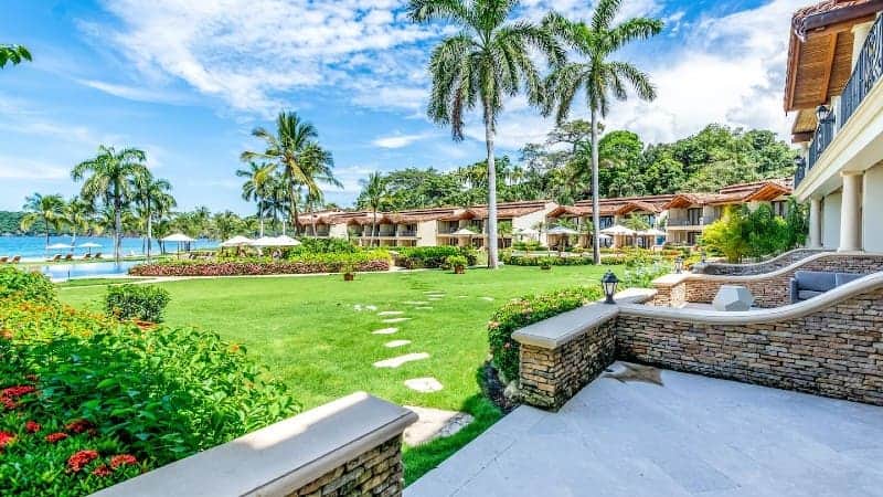 This image presents a view of a luxurious tropical resort. In the foreground, a manicured lawn is lined with stepping stones leading towards a collection of elegant bungalows with thatched roofs, nestled among tall palm trees. The bungalows overlook a tranquil body of water, likely a lagoon or beachfront, offering a picturesque view. The resort is lush with tropical vegetation, and bright flowering plants add splashes of color to the verdant landscape. 