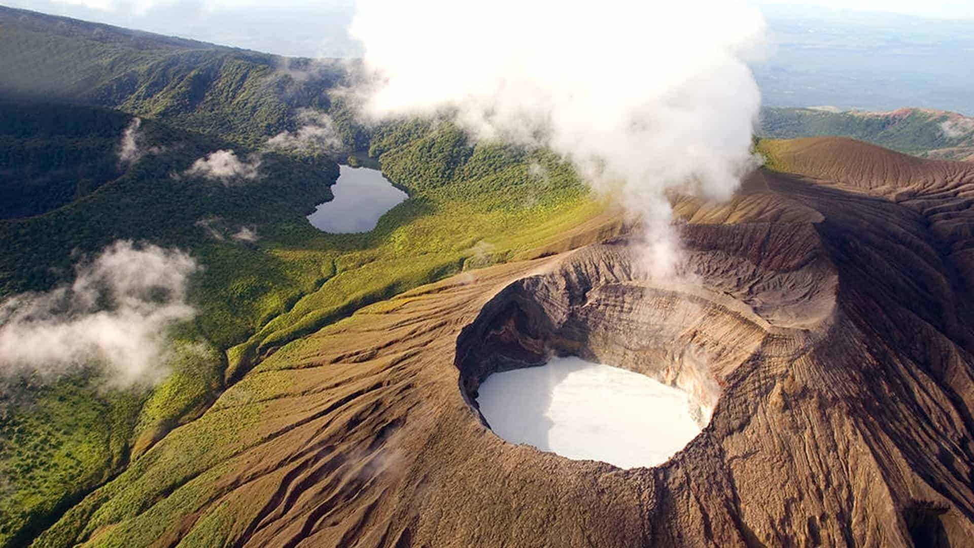 Aerial view of Rincon de la Vieja National Park, featuring the active crater with steam rising from it, a volcanic ash-covered slope, and lush greenery surrounding a calm lake in the volcanic basin.