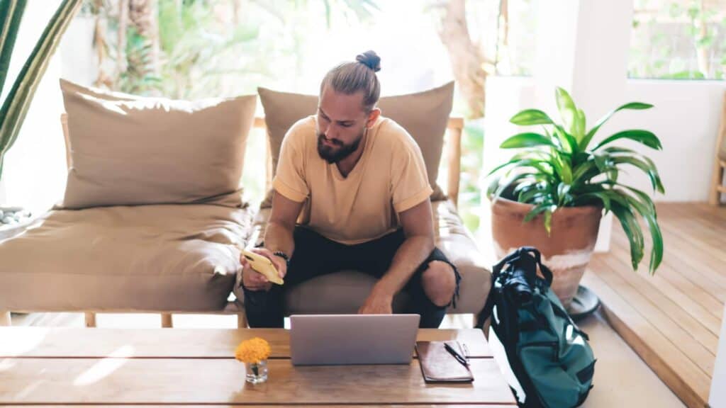 A man in a casual beige t-shirt and black trousers sits on a bench in a bright, airy room with a tropical feel, working on a laptop. He holds a phone in one hand, perhaps multitasking or taking a break. Nearby, a large potted plant adds a touch of greenery, and a comfortable daybed with pillows suggests a relaxed living space. On the wooden table, there's a small vase with yellow flowers, a notebook, and what appears to be a backpack on the floor next to the table, hinting at a mobile lifestyle or travel.