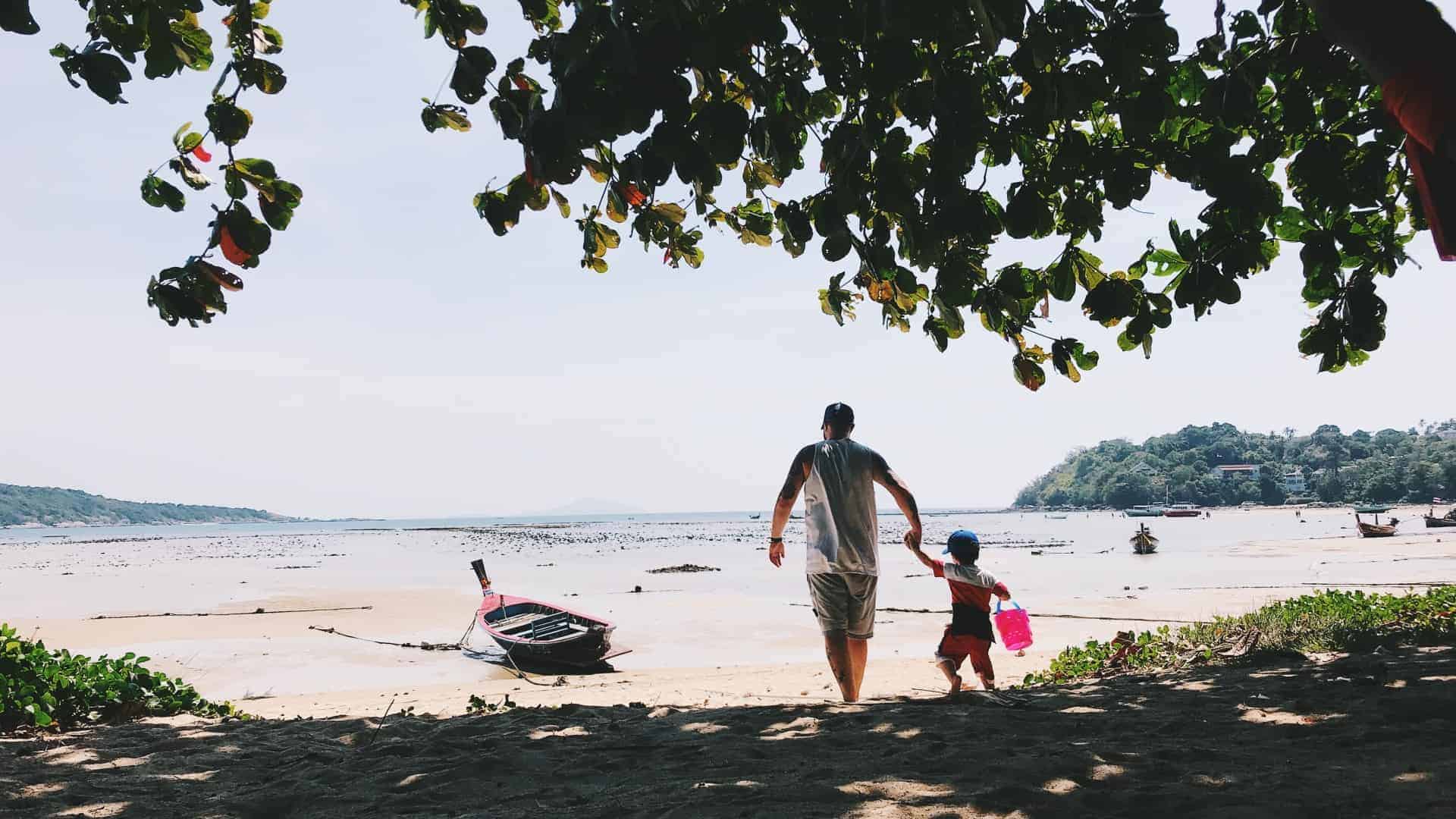 A serene beach scene with a man and a young child holding hands, walking towards the water's edge. The foreground shows a shaded area under the leafy canopy of a tree, while the background reveals a calm sea with a few boats anchored near the shore and hills in the distance. It is a bright, sunny day.