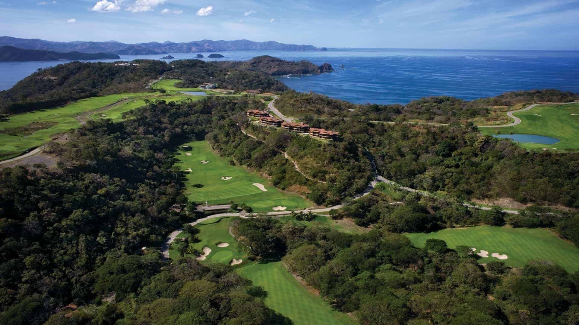 An aerial view of the Ocean Course at Peninsula Papagayo, coastal golf course with lush fairways, white sand bunkers, and multiple greens nestled among tropical forested hills. The course is adjacent to a deep blue ocean with scattered islands visible in the distance. Roads wind through the hills, and a row of terracotta-roofed structures sits atop one ridge, overlooking the scenic landscape.