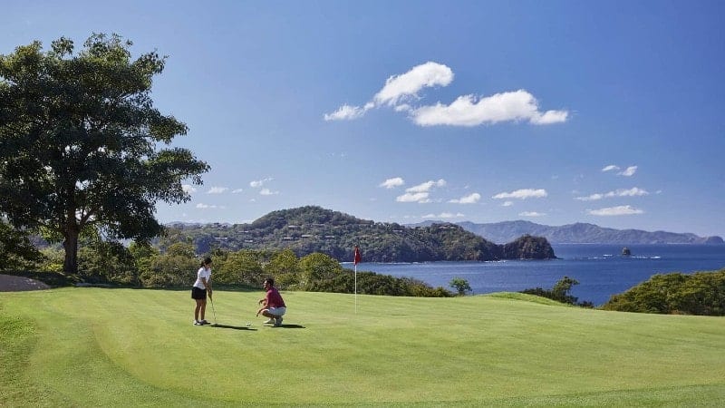 Two people on a lush green golf course, one standing and one crouching, likely discussing a shot. They are beside a hole with a red flag, under the shade of a tree. In the background, there's a panoramic view of a serene bay dotted with hills and islands under a clear blue sky with a few scattered clouds.