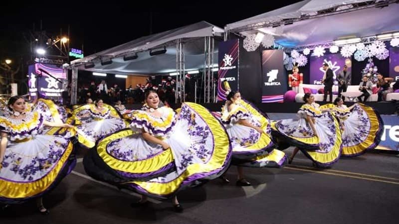 A group of dancers in traditional costumes perform at a nighttime cultural event. Their dresses have wide, swirling skirts in vibrant yellow and purple with decorative patterns, which they are twirling dramatically.