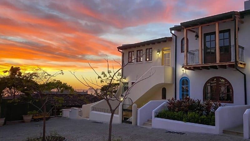 A picturesque view of a two-story house at sunset. The architecture features a mix of curves and arches, with white stucco walls and wooden accents around the windows. Warm lights glow from within and outside the house, complementing the vibrant orange and pink hues streaking across the sky. 