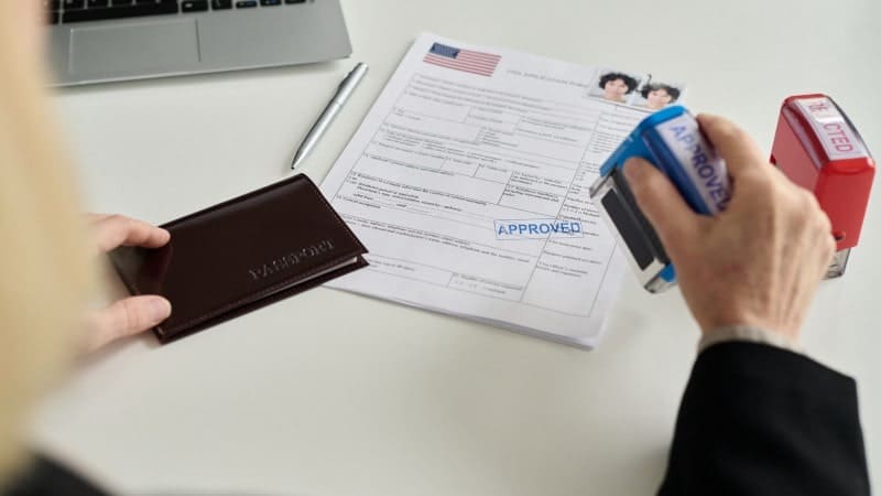 A person at a desk is about to stamp an 'APPROVED' stamp on a United States visa application form. There's a laptop, a silver pen, and a brown passport holder on the desk, indicating a professional setting likely within an immigration office.