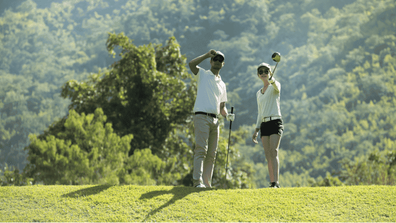 Two golfers are on a fairway with one golfer standing looking into the distance, shielding their eyes from the sun with a hand, and the other preparing for a tee shot with a driver in hand.
