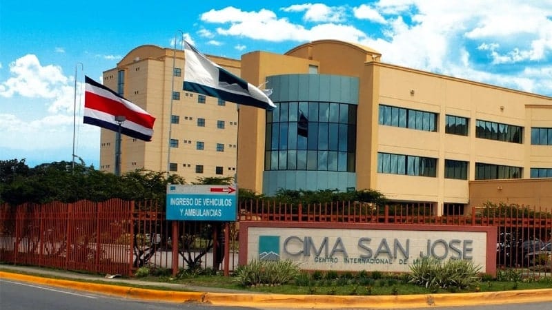 The front view of CIMA San José, a modern medical facility with a large glass facade and tan exterior walls. The Costa Rican flag and another flag are prominently displayed on flagpoles in front of the building. A red metal fence surrounds the property, and a sign indicating the entrance for vehicles and ambulances is visible at the foreground.
