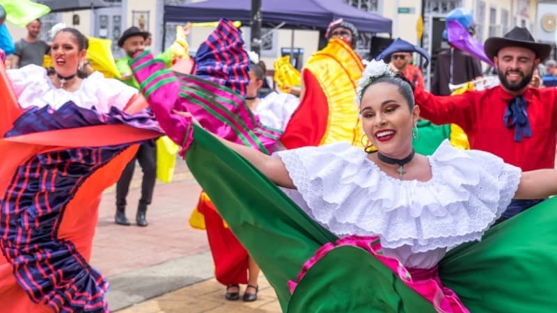 Dancers in colorful traditional attire perform in a daylight street festival. The central figure is a smiling woman with a flower in her hair, wearing a white blouse with ruffled sleeves and a vibrant green skirt with pink and red accents. Surrounding dancers have similarly bright, multicolored costumes with large, flowing skirts being twirled in the air.