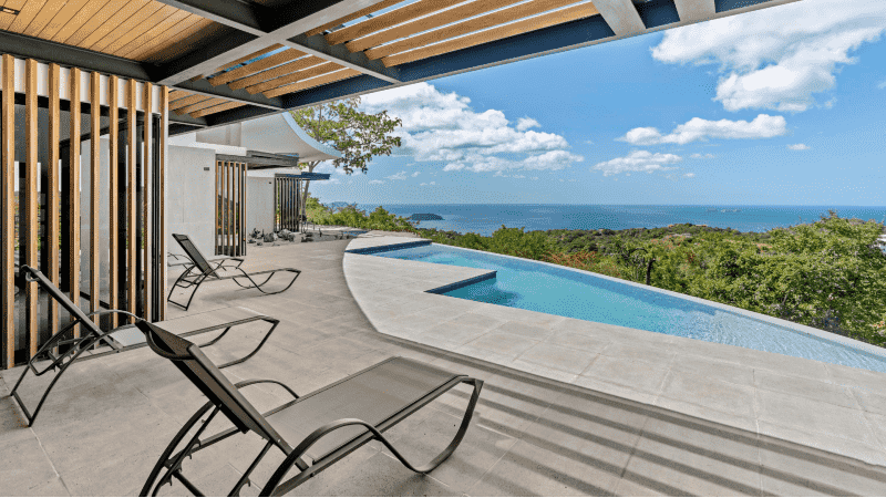 A luxurious infinity pool on a tiled patio with a stunning view of the ocean in the distance. There are lounge chairs on the left, and the modern architecture includes wooden slats as part of the roof design.