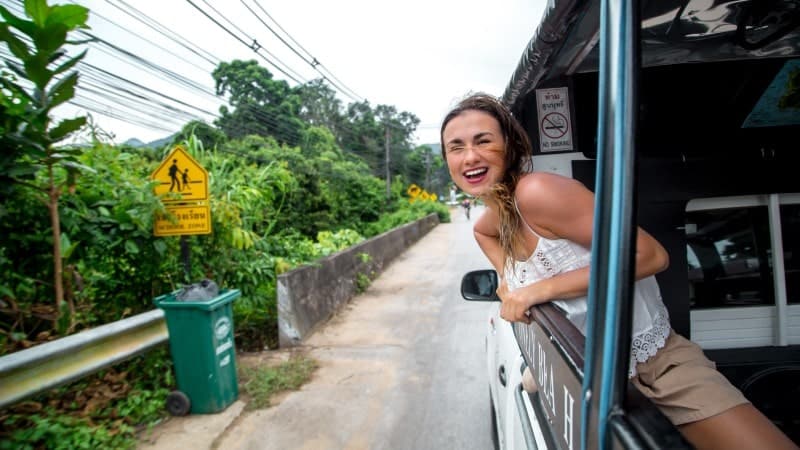 A joyful woman leaning out of the side of a tuk-tuk, her hair tousled by the wind, wearing a white sleeveless top and beige shorts. She is laughing and holding onto the vehicle, which is driving down a narrow road. The surroundings are lush and green with tropical vegetation. A yellow pedestrian crossing sign is visible in the background, along with a green trash bin by the roadside. 