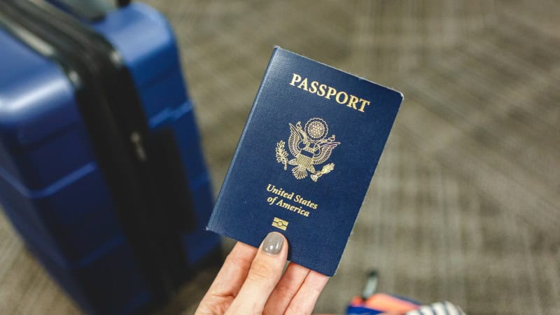 A hand holding a United States passport is in focus, with the iconic dark blue cover featuring the U.S. coat of arms. In the background, out of focus, is a blue suitcase, indicating a travel setting, possibly an airport or a travel hub.