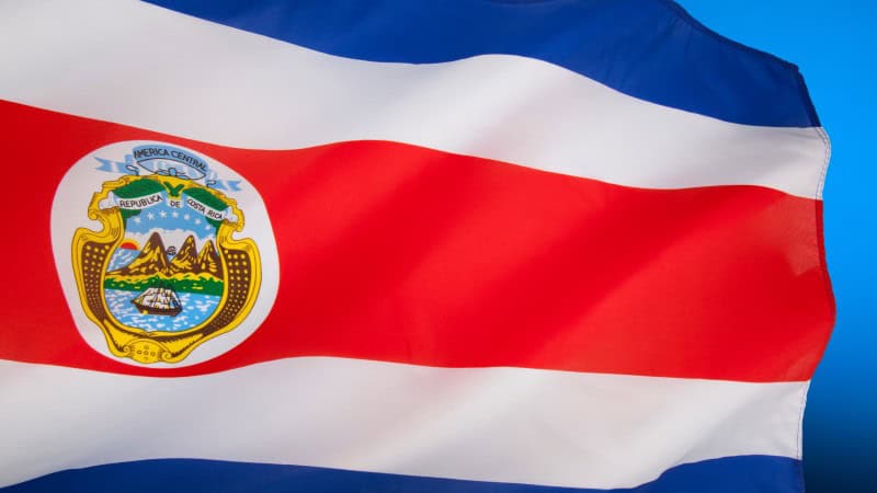 The flag of Costa Rica is unfurled and waving, with its vibrant horizontal bands of blue, white, and red, and the country's coat of arms prominently displayed in the white band.