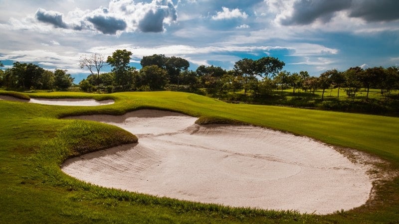 This image features a serene golf course with a large, pristine sand bunker in the foreground, curving into the lush green fairway. Beyond the bunker, the fairway is dotted with trees and undulating terrain under a dynamic sky with puffy white clouds and patches of blue.