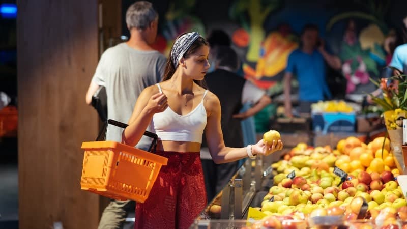 A young woman shopping at a fruit market, holding an orange shopping basket on her arm.