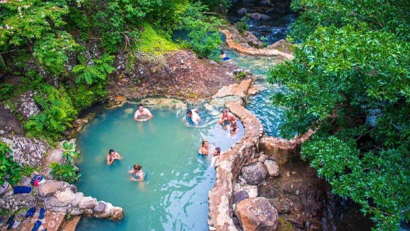 An aerial view of the Rio Negro Hot Springs where several people are enjoying the warm thermal waters in natural stone pools surrounded by lush tropical foliage.