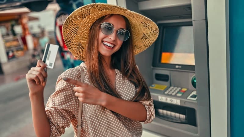 A joyful woman wearing a wide-brimmed straw hat and sunglasses stands in front of an ATM and is holding a credit card.
