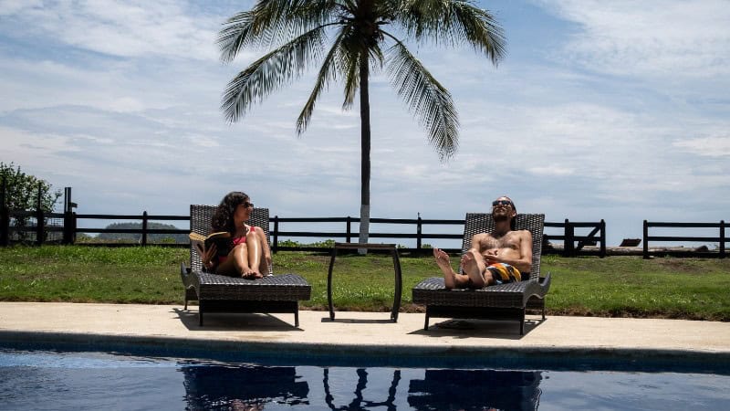 Two people are relaxing on sun loungers by a serene poolside that overlooks a tropical seascape. 