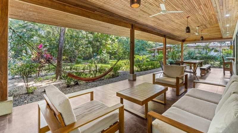 A serene outdoor patio area seamlessly blending with a lush tropical garden. The patio is sheltered with a wooden slatted roof and equipped with modern wooden lounge furniture with comfortable beige cushions.