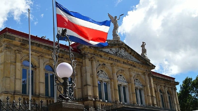 The National Theater of Costa Rica on a bright day, with the Costa Rican flag proudly waving in the wind. The classical architecture of the building features ornate statues, an elegant facade with multiple arch-shaped windows, and a pediment topped with a sculptural group. An ornamental street lamp is visible in the foreground, and the sky behind is partly cloudy, casting a soft light on the theater’s historical structure.