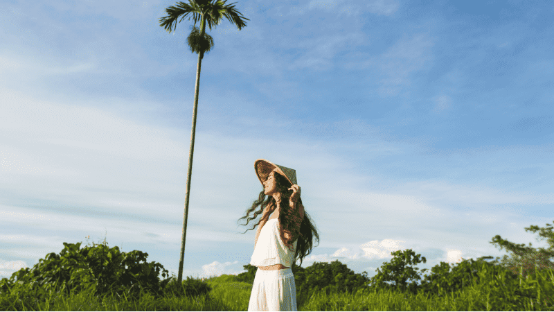 A woman in a white dress and a straw hat standing in a lush green field, her gaze directed upward. She is holding the hat against a gentle breeze, which also stirs her long, wavy hair. In the background, a tall palm tree stretches into the sky.