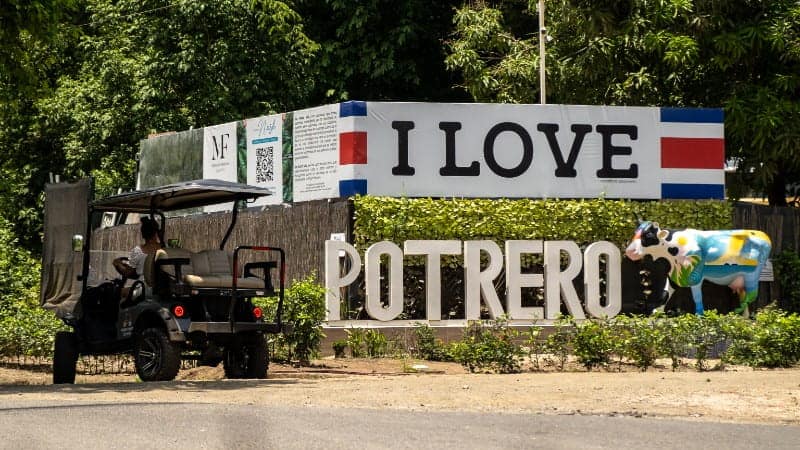 A large sign reading "I LOVE POTRERO" is prominently displayed at the roadside, with each letter standing independently on a base and painted in the colors of the Costa Rican flag. Next to the sign, there's a colorful painted cow statue.