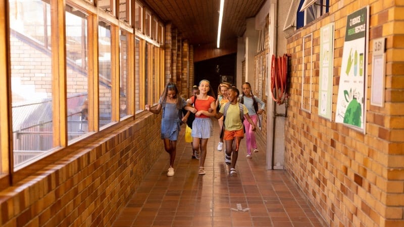 A group of cheerful young students walking down a school hallway, bathed in warm sunlight from the windows on the left. The girls are diverse in appearance, chatting and smiling, carrying colorful backpacks, and the hallway is lined with educational posters on the right-hand side, adding to the atmosphere of a lively educational environment.