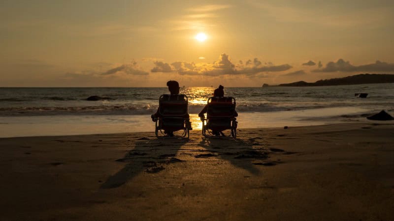 Two people in silhouette sit side by side in beach chairs on the shore, facing the ocean as the sun sets on the horizon. The sky is painted with warm hues of orange and yellow, reflecting on the water's surface, while gentle waves lap onto the sand.