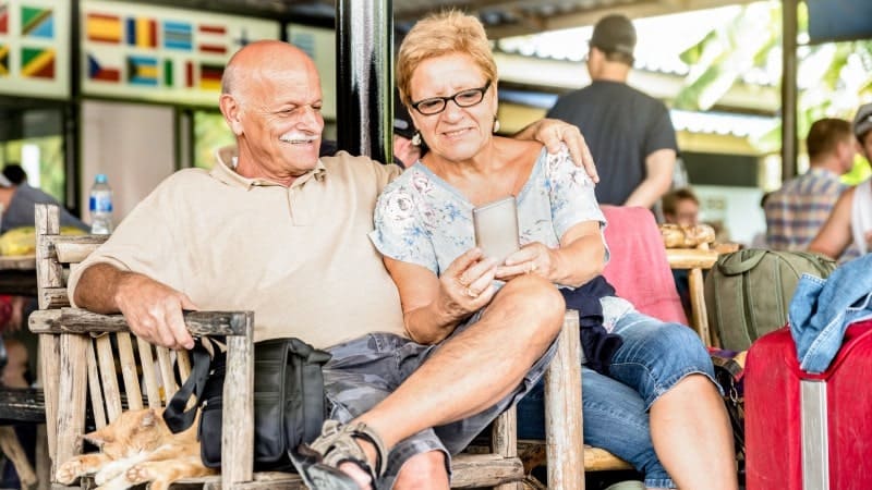 An elderly couple sits closely together on a wooden bench, smiling and looking at a smartphone screen. The man has his arm around the woman.